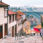 Narrow stone steps between colorful houses with red tiled roofs leading toward a blue arched bridge over a river