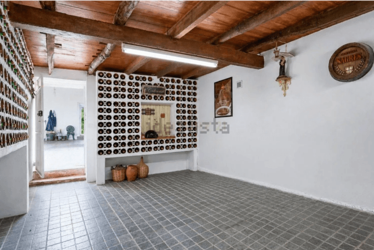 Wine room with wall mounted racks holding hundreds of bottles around a central pass through window and a tiled floor under wooden beams on the ceiling