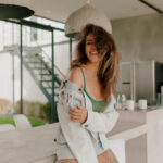 Smiling woman in a light denim jacket and green top leaning on a marble counter in a bright modern kitchendining area