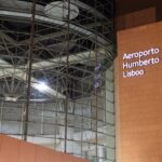 Interior of an airport terminal with exposed steel beams and an orange wall displaying Aeroporto Humberto Delgado Lisboa