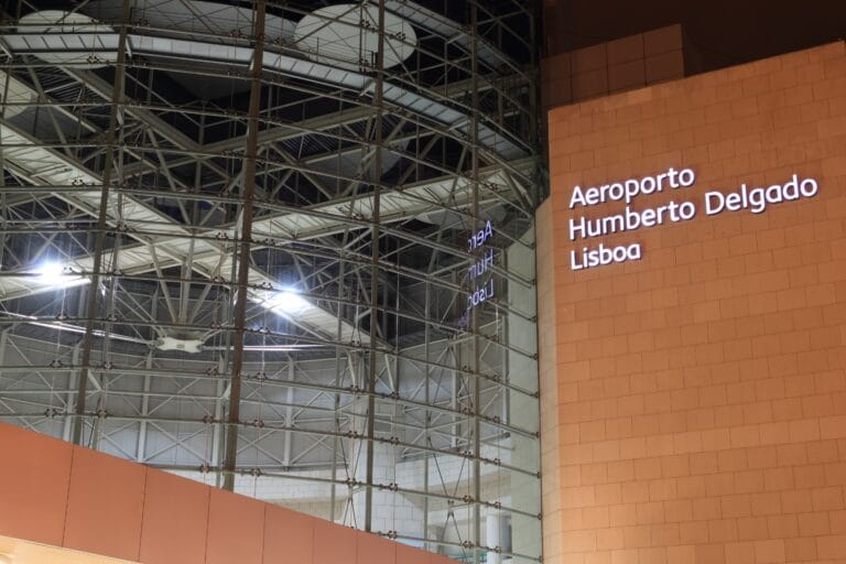 Interior of an airport terminal with exposed steel beams and an orange wall displaying Aeroporto Humberto Delgado Lisboa
