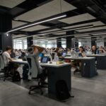 Open plan tech workspace with rows of desks and people working on laptops under long overhead lights