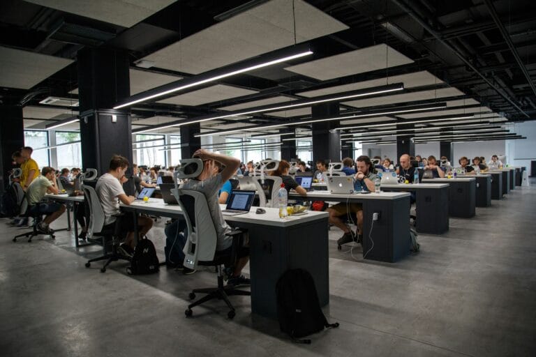Open plan tech workspace with rows of desks and people working on laptops under long overhead lights
