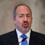 Close up of a man in a dark suit and blue tie speaking at a podium during a press briefing with a blurred flag in the background