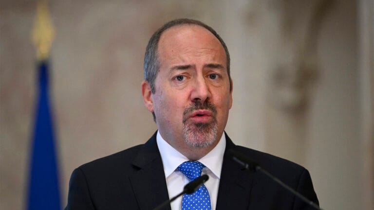 Close up of a man in a dark suit and blue tie speaking at a podium during a press briefing with a blurred flag in the background