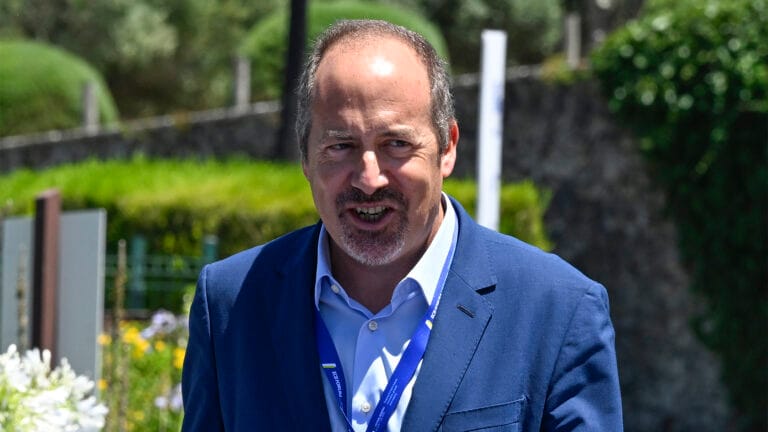 Man in a blue suit smiling outdoors at an event wearing a lanyard with a badge