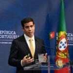 Man in a dark suit and yellow tie speaks at a podium with the Portuguese flag and EU flag in the background
