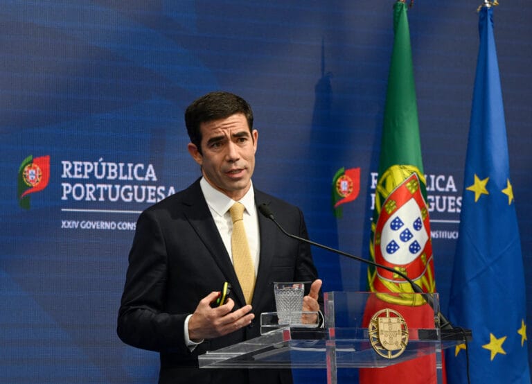 Man in a dark suit and yellow tie speaks at a podium with the Portuguese flag and EU flag in the background