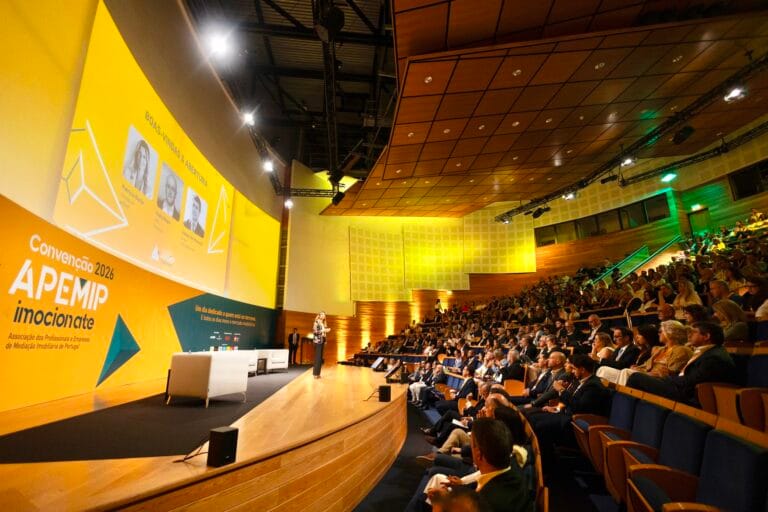 Speaker on stage in a bright yellow conference backdrop addressing a large audience in a tiered auditorium