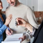 Couple smiles as a real estate agent hands over house keys while they review a contract in the background