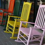 Colorful wooden rocking chairs in a row outside a storefront from red to pink along a glass window