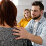 Man with a beard sits facing a woman resting his hand on her shoulder in a counseling or supportive conversation another woman in a yellow top observes in the background in a bright modern office