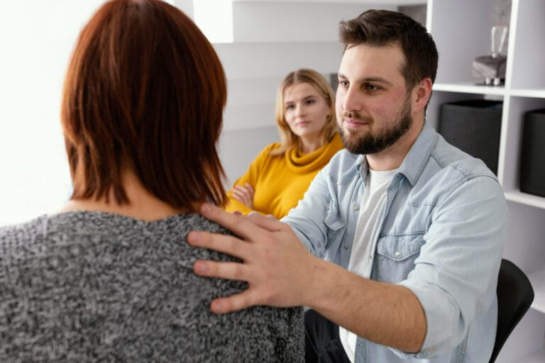Man with a beard sits facing a woman resting his hand on her shoulder in a counseling or supportive conversation another woman in a yellow top observes in the background in a bright modern office