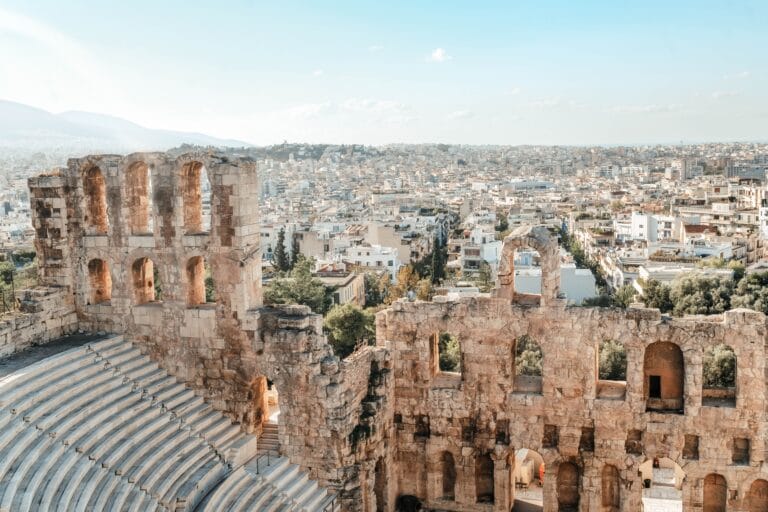 Ancient stone amphitheater arches overlook a modern city spread across the hills under a clear blue sky