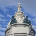 Dome of Banco de Portugal with two seated green statues atop a decorative white column against a blue sky