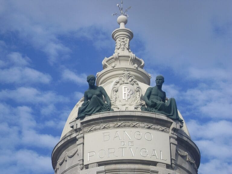 Dome of Banco de Portugal with two seated green statues atop a decorative white column against a blue sky
