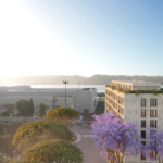 Sunlit urban street with modern beige buildings and purple flowering trees sea visible in the distance under a clear blue sky