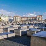 Field of uniform concrete blocks of varying heights in a grid at the Berlin Holocaust Memorial with buildings in the background and a clear blue sky