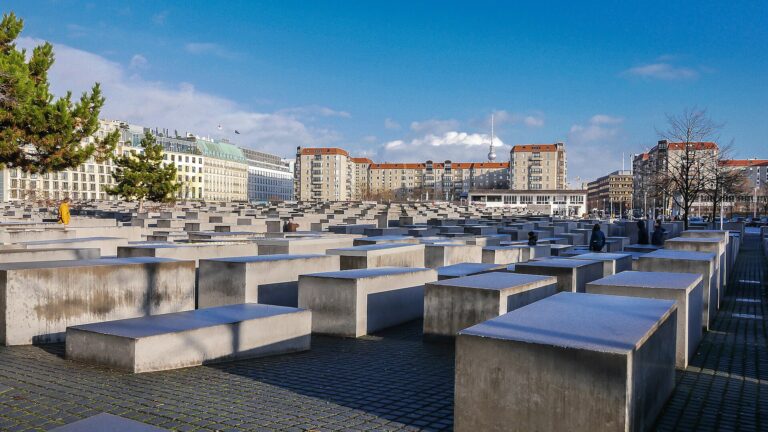 Field of uniform concrete blocks of varying heights in a grid at the Berlin Holocaust Memorial with buildings in the background and a clear blue sky