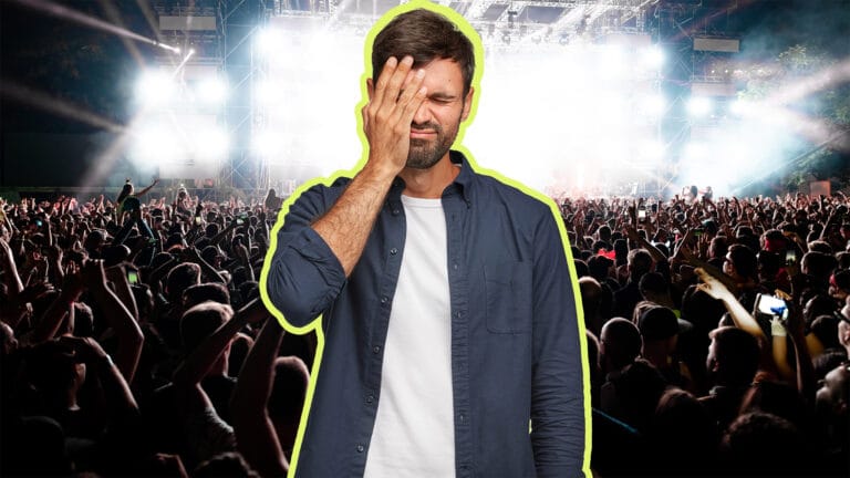 Man in a casual blue shirt and white tee covers one eye with his hand at a packed night concert crowd in the background