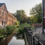Brick buildings line a calm canal with reflections while an outdoor seating area with tables and chairs runs along a railing on the right