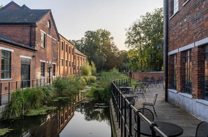 Brick buildings line a calm canal with reflections while an outdoor seating area with tables and chairs runs along a railing on the right