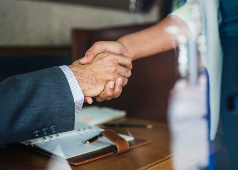 Close up of two professionals shaking hands over a wooden desk with a binder and notebook in the foreground signaling an agreement