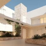 Modern white apartment complex with numbered doors along a sunlit courtyard and stone planters with greenery behind a low wall