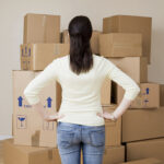 Back view of a woman with dark hair tied in a ponytail hands on hips standing among stacked cardboard boxes in a room