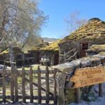Rustic stone cottage with a mossy roof and a wooden picket gate in a sunny garden setting with bare trees nearby