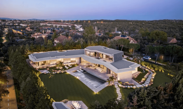 A modern L shaped mansion with a central pool and lush lawn viewed from above at dusk