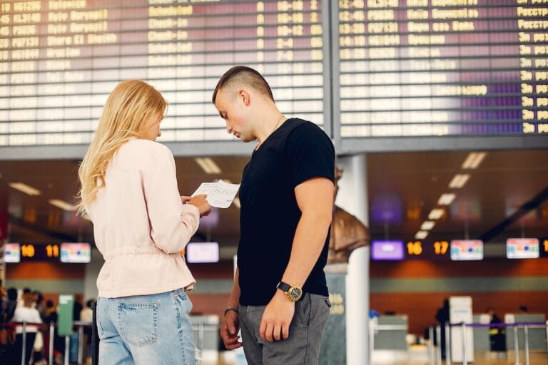 Couple at an airport check in area looking over a boarding pass with a large flight information board overhead