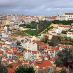 A panoramic view of a walled hillside city with colorful red roofed buildings and a church atop a hill surrounded by trees and green terraces