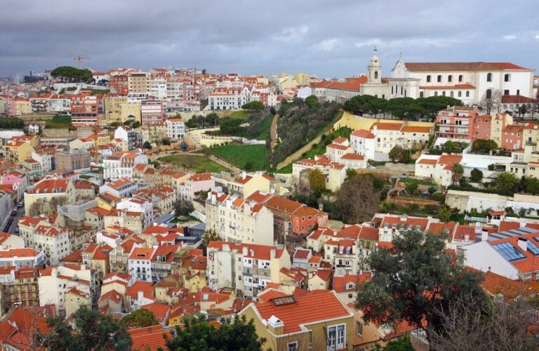 A panoramic view of a walled hillside city with colorful red roofed buildings and a church atop a hill surrounded by trees and green terraces
