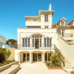Light colored multi story villa with arched doors a central balcony and a wide courtyard under a blue sky