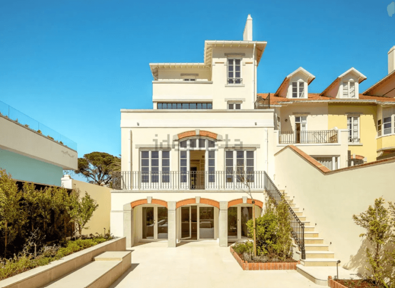 Light colored multi story villa with arched doors a central balcony and a wide courtyard under a blue sky