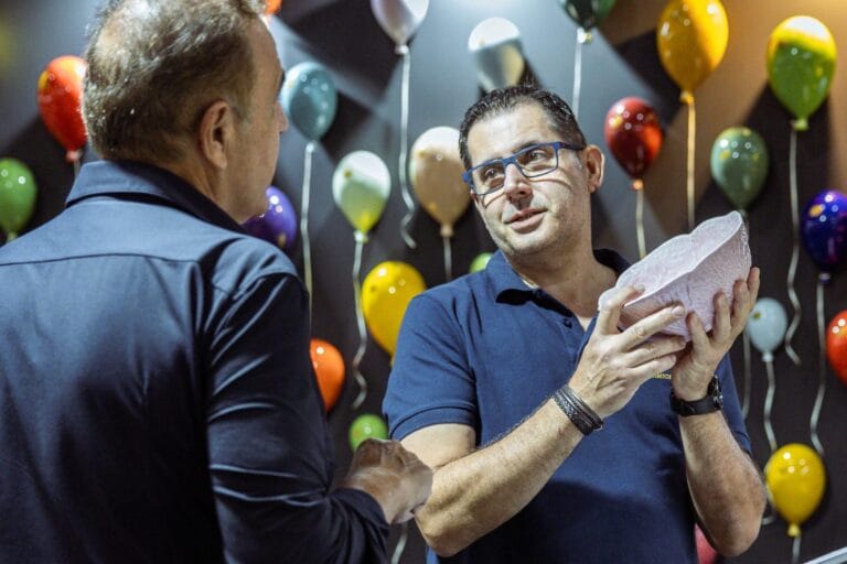 Two men in blue shirts chat at a party with colorful balloons in the background the man on the right holds a pink decorative item