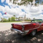 Rear view of a glossy red vintage convertible parked on a street with a Cuban flag and a statue in the background beneath a blue sky with clouds