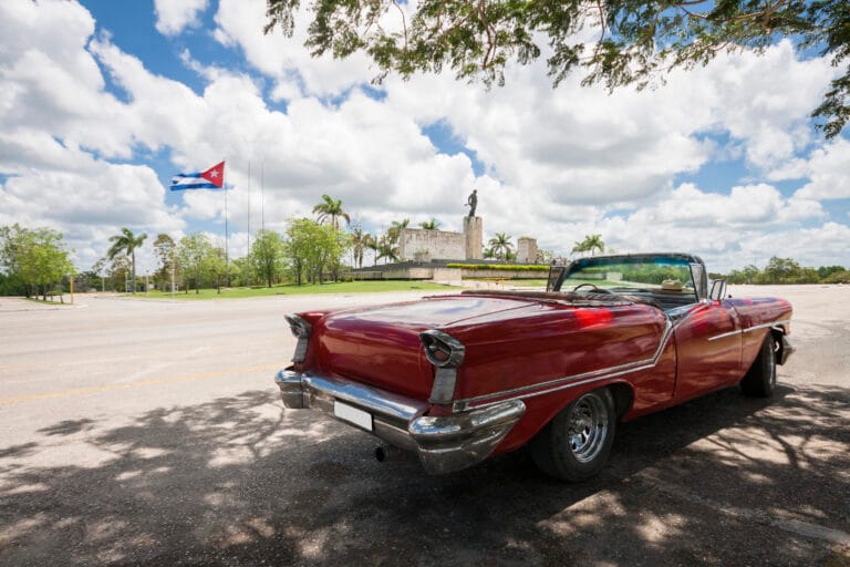 Rear view of a glossy red vintage convertible parked on a street with a Cuban flag and a statue in the background beneath a blue sky with clouds