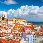 Colorful hillside coastal town with pastel houses and red tiled roofs a white domed church tower rises above the buildings against a blue sky and clouds