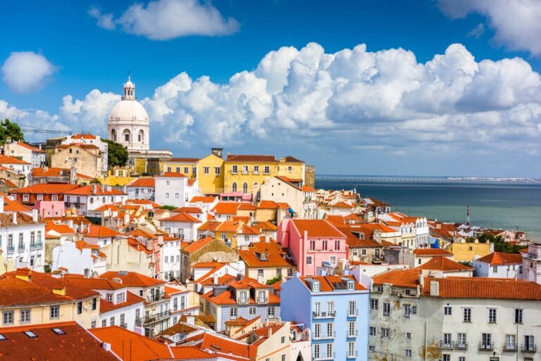 Colorful hillside coastal town with pastel houses and red tiled roofs a white domed church tower rises above the buildings against a blue sky and clouds