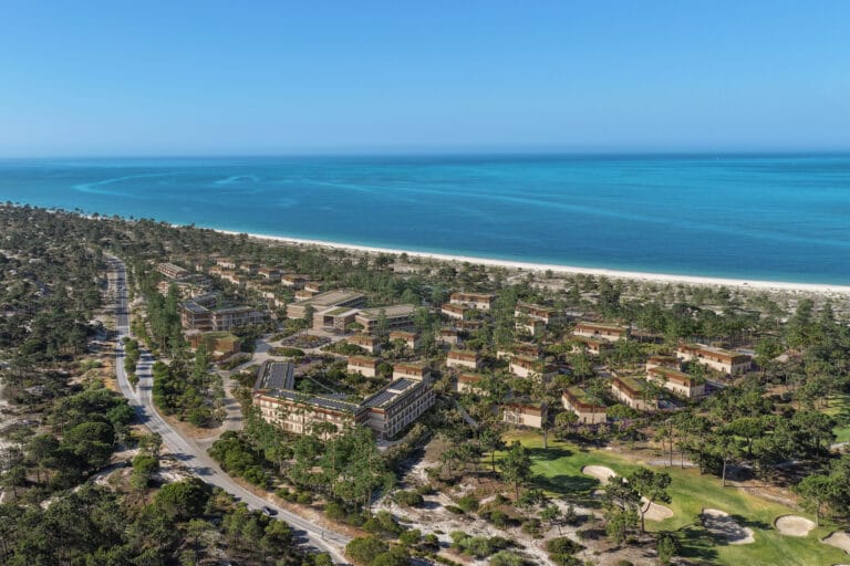 Aerial view of a coastal resort town with a sandy beach turquoise sea and pine forested hills surrounding low rise buildings and roads