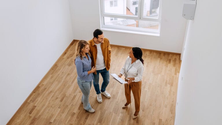 Three adults in a bright empty apartment discuss plans one holding a clipboard and smiling