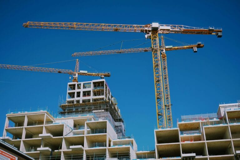 Construction site with tall yellow cranes above unfinished concrete apartment blocks against a clear blue sky