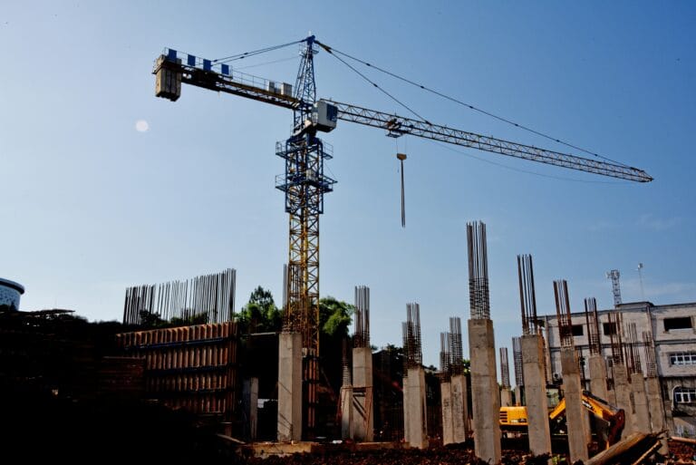 Tower crane at a construction site with concrete columns and exposed steel rebar against a clear blue sky indicating ongoing building work