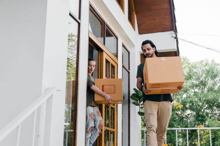 Two people carry cardboard boxes on a modern homes porch preparing to move in or out man in foreground with a large box labeled TOYS and woman at the doorway behind him