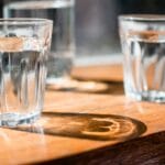Two clear glass tumblers of water on a sunlit wooden table with reflections and shadows