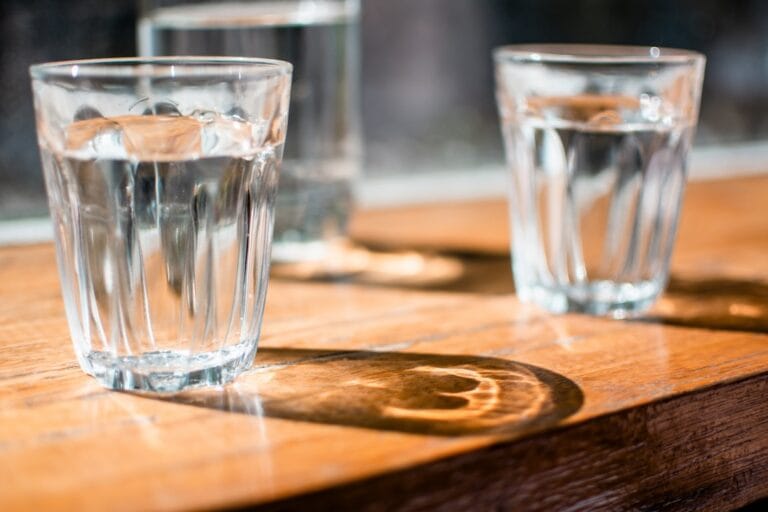 Two clear glass tumblers of water on a sunlit wooden table with reflections and shadows