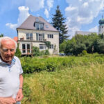 Older man in a white polo standing in a grassy yard with a large house and a church tower with a green onion dome in the background on a sunny day