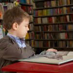 Young boy in a brown jacket reads a large open book at a red table in a library
